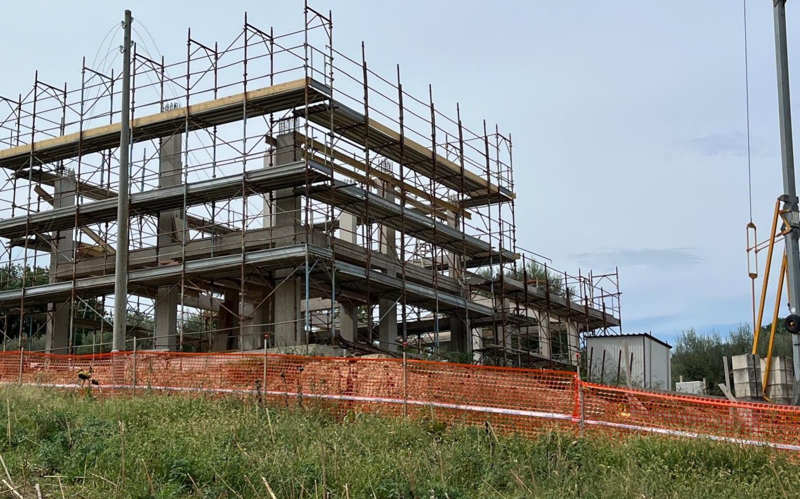 La struttura vista da fuori, coi ponteggi su tutti i fronti e la recinzione di cantiere arancione.