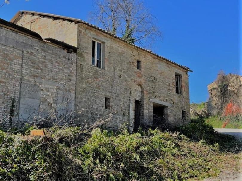 Il fronte lungo in pietra prima dei lavori, con le finestre vuote e la vegetazione tagliata ammucchiata davanti.
