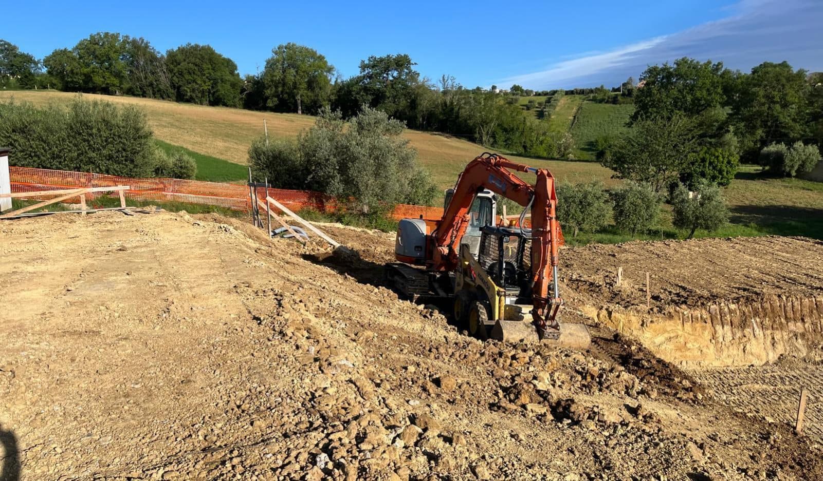 Lo scavo delle nuove fondazioni sul pendio, con l’escavatore al lavoro e le colline di Servigliano dietro.